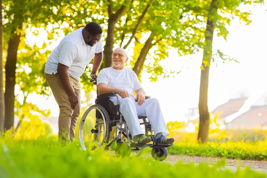 Caregiver pushing a smiling older man in a wheelchair through a sunny park