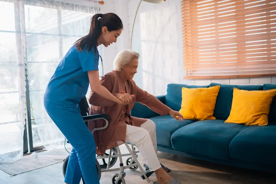 Caregiver helping an older woman stand from her wheelchair in a living room