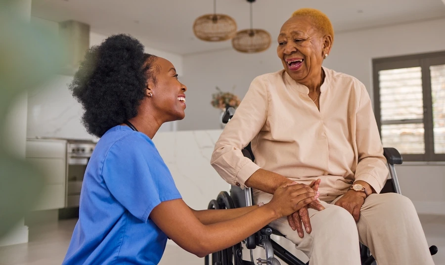 Nurse kneeling and holding hands with a laughing older woman in a wheelchair