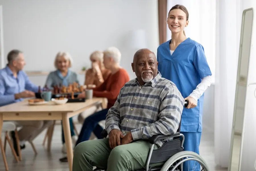Smiling nurse beside an older man in a wheelchair, with residents at a table behind