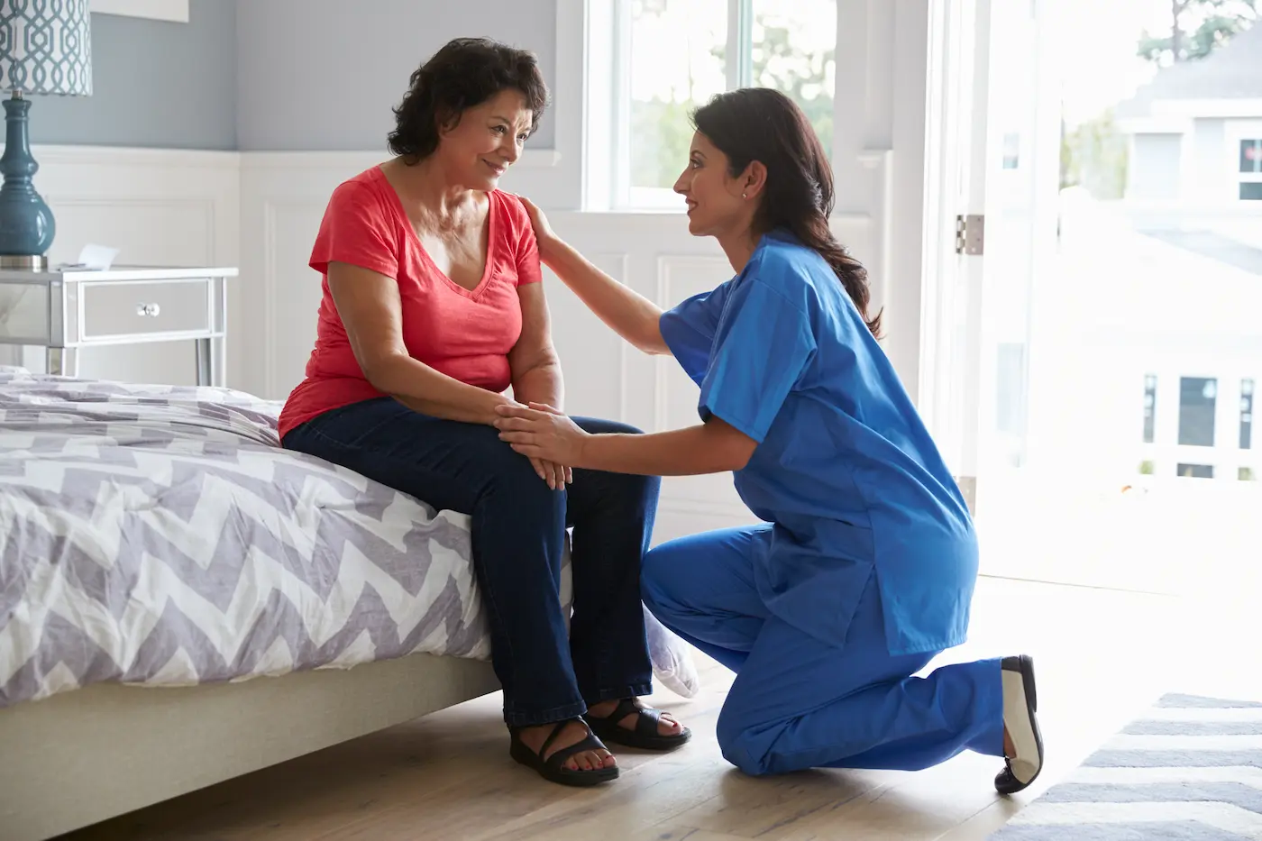 Nurse kneeling to talk with a woman sitting on her bed