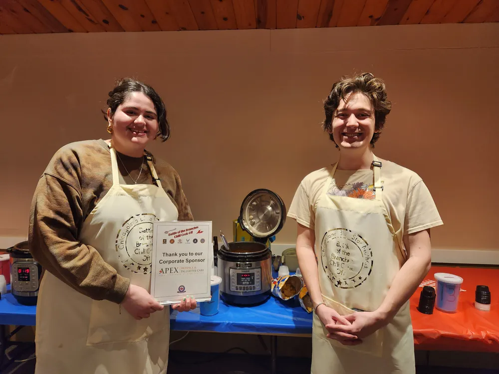 Dos voluntarios con delantales junto a ollas de cocción lenta en un concurso de chili.