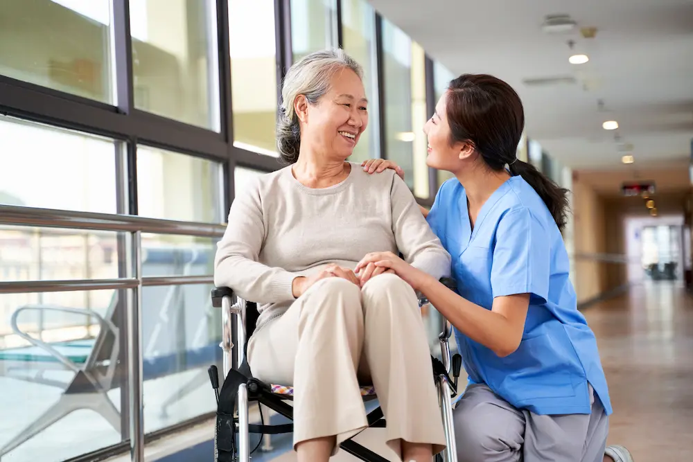 Nurse kneeling beside an older woman in a wheelchair in a hallway