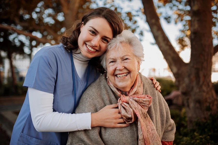 Una cuidadora abraza a una anciana sonriente al aire libre.