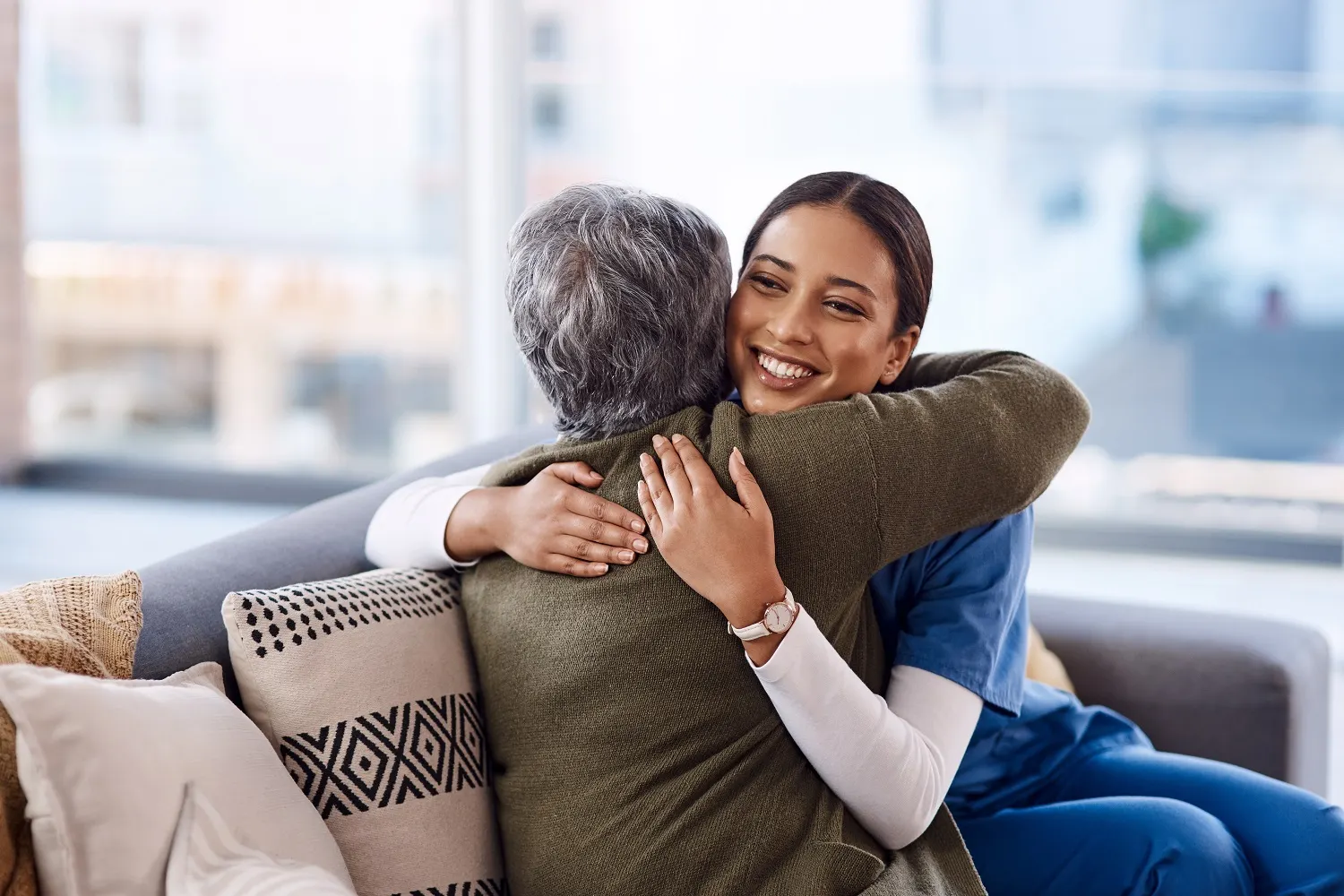 Una cuidadora sonriente, vestida con uniforme médico, abraza a una anciana en un sofá.
