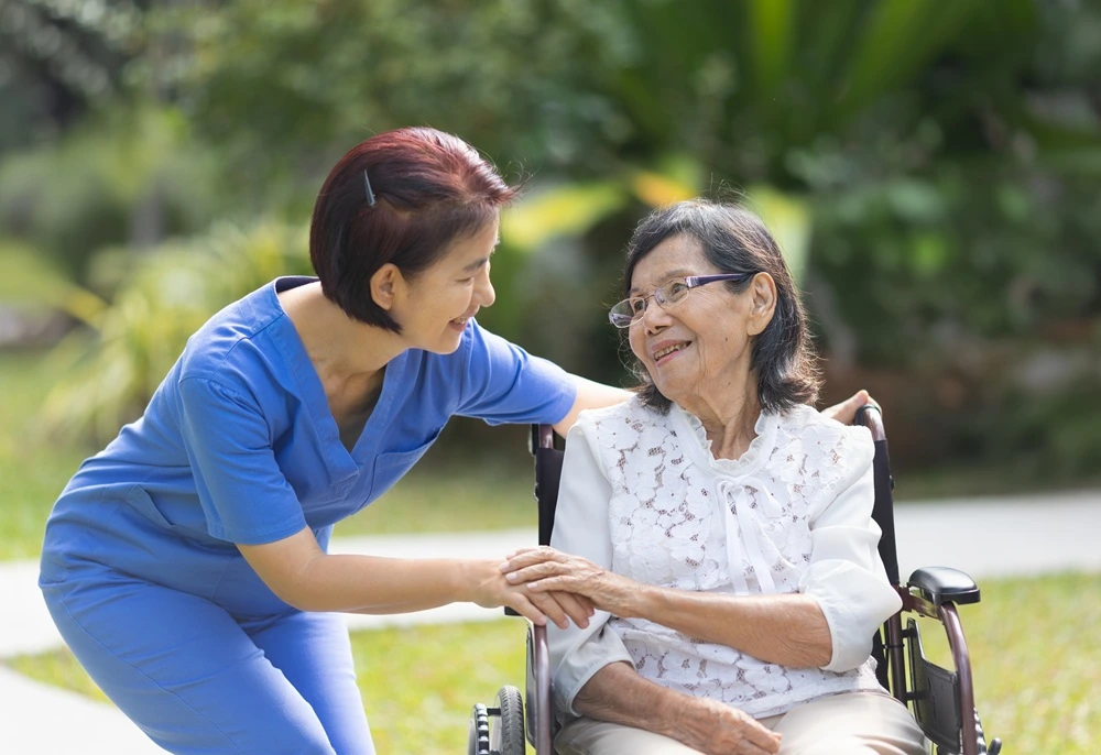 Una cuidadora se arrodilla junto a una anciana sonriente en silla de ruedas al aire libre.