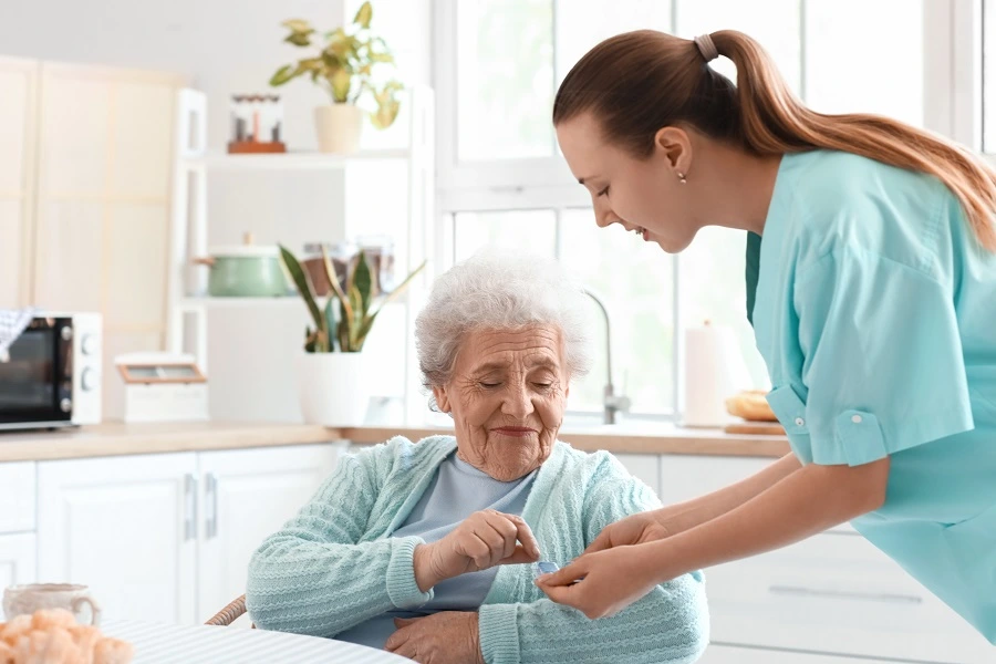 Caregiver in scrubs helping an older woman button her cardigan in a kitchen