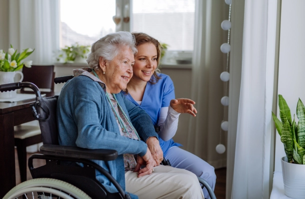 Caregiver sitting beside an older woman in a wheelchair by a window