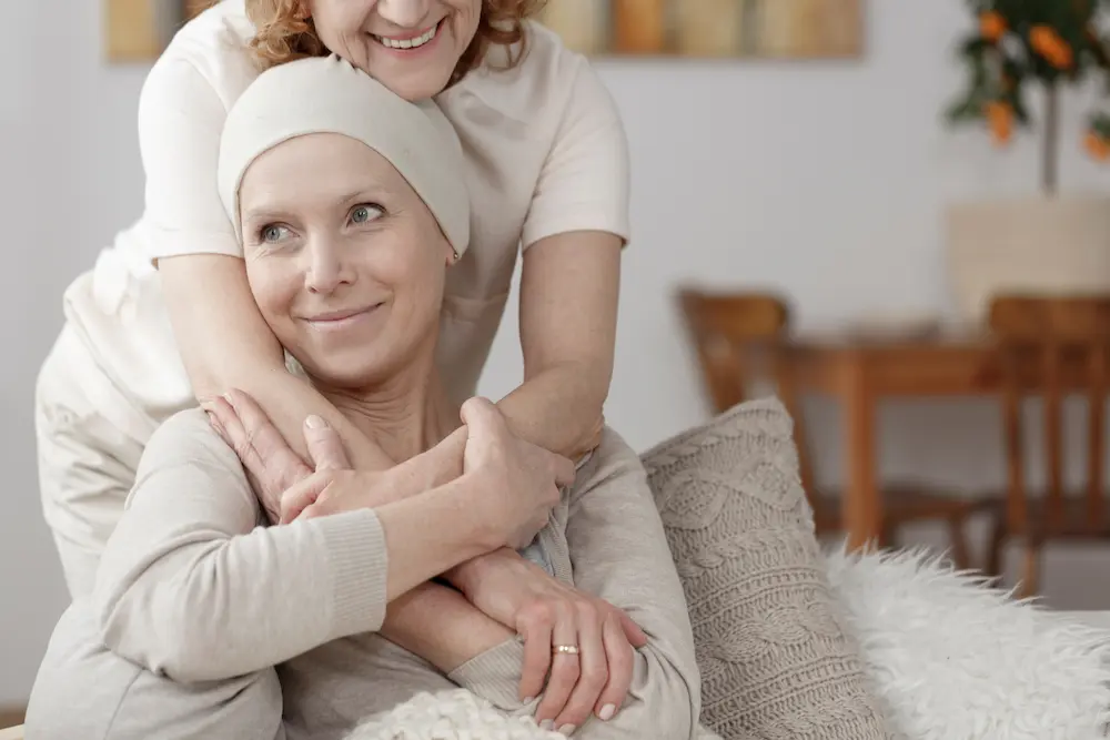 Mujer abraza a una paciente de cáncer sonriente que lleva un pañuelo en la cabeza.
